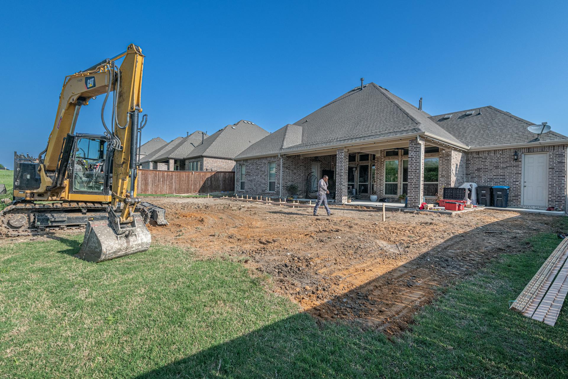 Residential back yard stripped back to bare soil beside remaining lawn, with a small tracked excavator working the ground